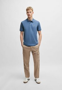 Niebieska koszulka polo z kołnierzykiem i logo, w połączeniu z beżowymi spodniami cargo z bocznymi kieszeniami. Model nosi białe sneakersy.