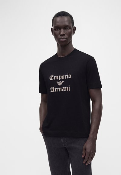 Μαύρος αρσενικός μοντέλο που φοράει μαύρο T-shirt Emporio Armani με μπεζ λογότυπο, στέκεται μπροστά σε απλό ανοιχτό γκρι φόντο.