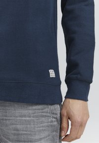 Marineblauwe sweatshirt met een getextureerde stof, voorzien van een klein wit label met tekst. Grijze spijkerbroek gedeeltelijk zichtbaar aan de onderkant.