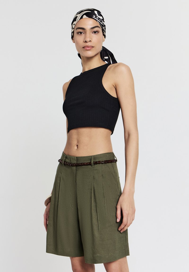 Donna che indossa un top corto senza maniche nero, pantaloni corti plissettati verde oliva con una cintura intrecciata e un foulard a motivi bianco e nero.