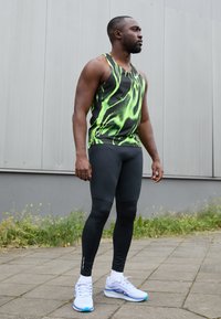 Abbigliamento sportivo composto da una maglietta senza maniche nera con un motivo astratto verde, leggings neri aderenti e scarpe bianche con accenti blu.