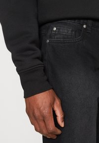 Mano de piel oscura descansando al lado de unos jeans de mezclilla negros con un bolsillo frontal visible y un puño de camisa de manga larga negra.