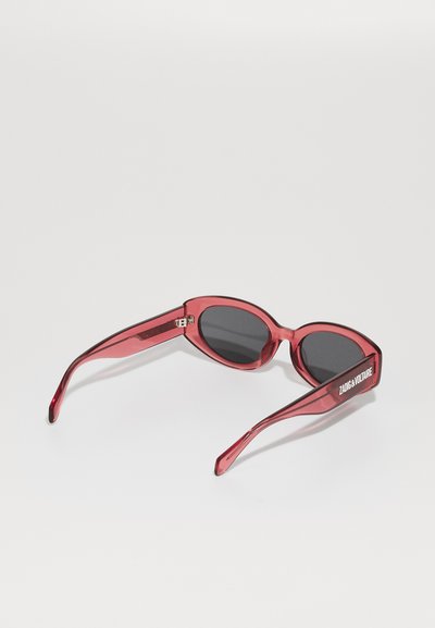 Lunettes de soleil rouges transparentes avec des verres ovales, des branches fines et des détails texturés. Marquées "ZADIG & VOLTAIRE" sur le côté.
