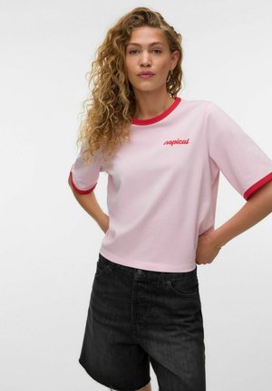 Kobieta o kręconych włosach, ubrana w jasnoróżowy t-shirt z czerwonym wykończeniem i napisem "tropical", w połączeniu z czarnymi jeansowymi szortami, pozująca z rękami na biodrach.