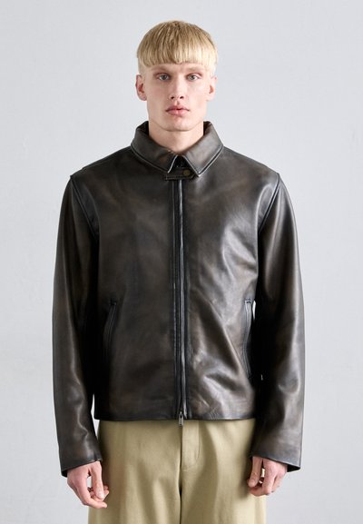 Brown leather jacket with a high collar, zip front, side pockets, and a smooth texture. Model wears loose-fitting beige trousers.