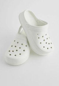 Sabots blancs avec des trous de ventilation, fabriqués en matériau léger. Présentent un embout arrondi et une sangle arrière pour un ajustement sécurisé. Texture lisse.