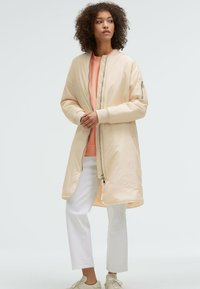 Hellbeige Bomberjacke mit Rei ßverschluss vorne, gerippten Bündchen und gefüttertem Innenfutter. Kombiniert mit einem pfirsichfarbenen Sweatshirt und weißen Hosen. Glatte Textur.