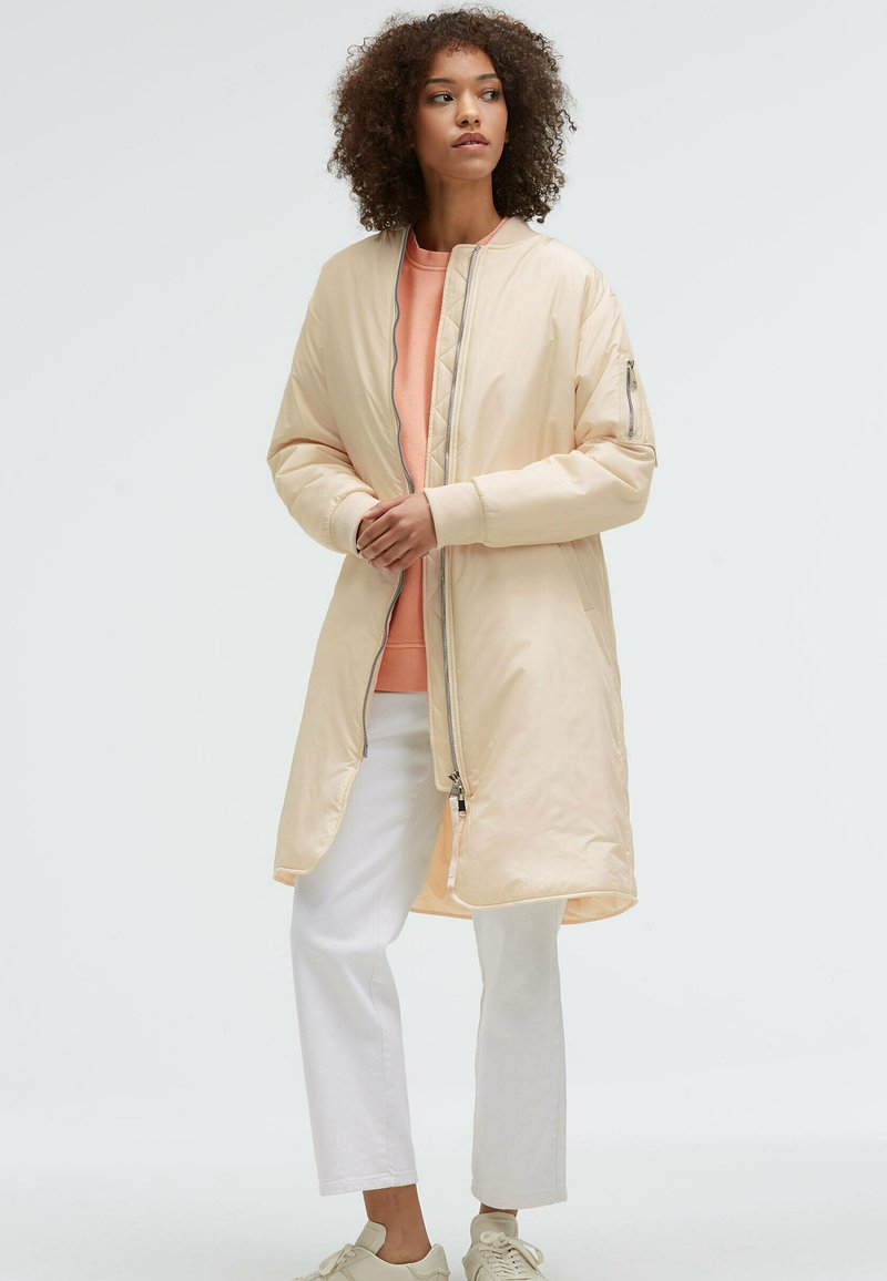 Hellbeige Bomberjacke mit Reißverschluss vorne, gerippten Bündchen und gefüttertem Innenfutter. Kombiniert mit einem pfirsichfarbenen Sweatshirt und weißen Hosen. Glatte Textur.