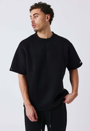Jonge man met krullend haar die een zwarte oversized t-shirt en zwarte broek draagt, kijkt naar links met één hand in zijn zak.