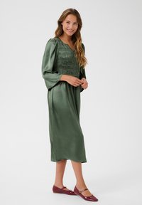 Green satin dress with a smocked bodice, long sleeves, and a flowing skirt; paired with burgundy flats featuring a strap detail.