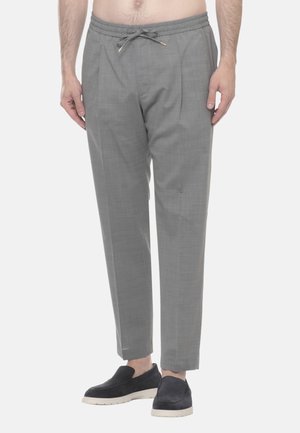 Uomo che indossa pantaloni grigio chiaro con coulisse e scarpe nere slip-on, in piedi davanti a uno sfondo chiaro e uniforme.