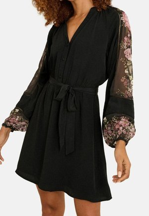 Donna che indossa un vestito nero con maniche lunghe trasparenti con motivo floreale e cintura annodata in vita.