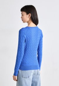 Suéter azul de punto trenzado con dobladillos acanalados, cuello redondo y mangas largas. Combinado con jeans de un azul claro, mostrando una superficie texturizada.