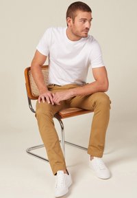 Weißes T-Shirt, beige Chinos, weiße Sneakers. Lässiger Schnitt, glatte Textur, modernes Design. Modell sitzt auf einem Holzstuhl mit Rattan-Elementen.