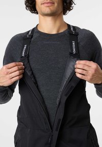 Black insulated overalls with adjustable straps, front zip closure, and side pockets. Worn over a dark gray long-sleeve shirt.