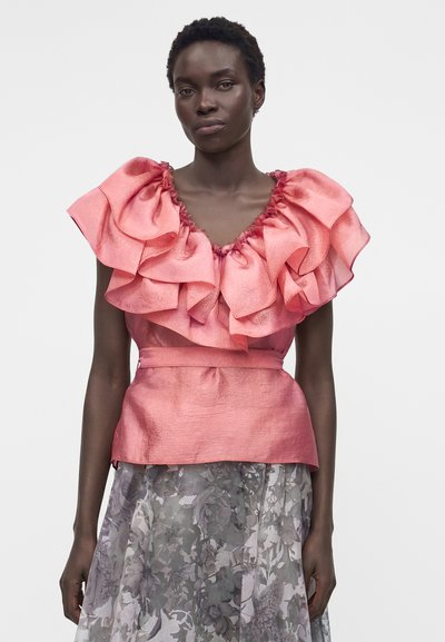 Γυναίκα με ροζ τοπ με βολάν και εφαρμοστή μέση, φορούν μια διάφανη γκρι φλοράλ φούστα, στέκεται σε λευκό φόντο.