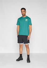 Τιρκουάζ βαμβακερό t-shirt με λογότυπο σε διαβάθμιση χρωμάτων, συνδυασμένο με μαύρο αθλητικό σορτς. Το μοντέλο φορά μαύρα αθλητικά παπούτσια και καλτσάκια μέχρι τον αστράγαλο.