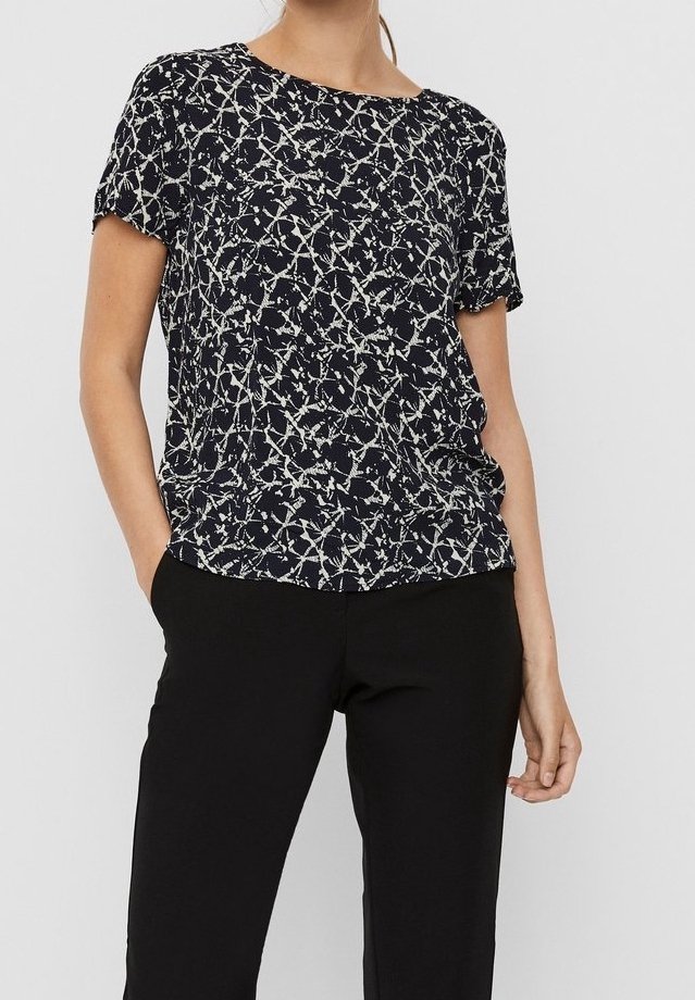 Donna con un top corto a maniche corte a righe astratte in bianco e nero, inserito nei pantaloni neri, in piedi con una mano in tasca.