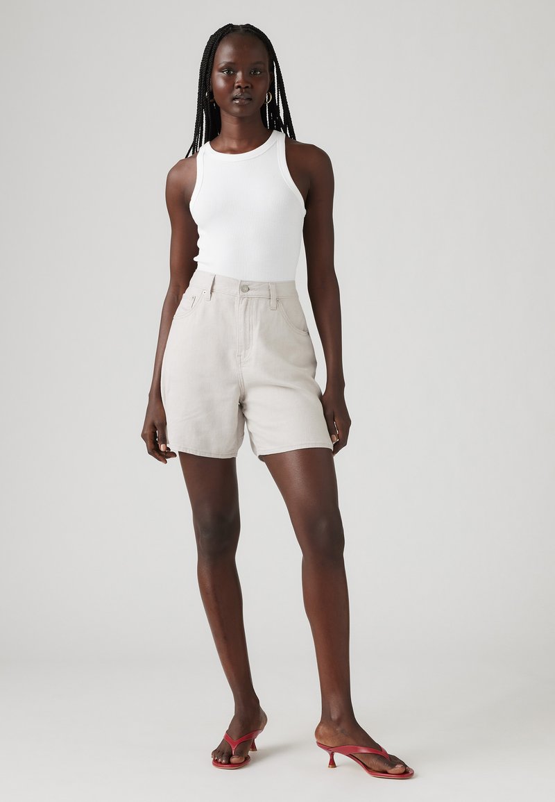 Helle beige High-Waisted-Shorts aus Baumwollgemisch, mit einem Frontknopfverschluss, vier Taschen und glatter Oberfläche. Kombiniert mit einem weißen Tanktop und roten Flip-Flops.