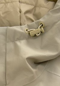 Lys beige jakke med krave, lavet af glat, struktureret stof; inkluderer en trækkontakt med "SIGNATURE" prægede på. Syningerne er synlige.