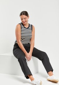 Top sin mangas a rayas en azul marino y crema, combinado con pantalones negros. Modelo sentada en un cubo blanco, vistiendo zapatillas blancas con suelas de yute.