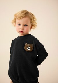 Sweat-shirt noir avec un patch de visage de ours brun rond sur la poitrine gauche. Tissu doux, coupe décontractée, poignets et ourlet côtelés.