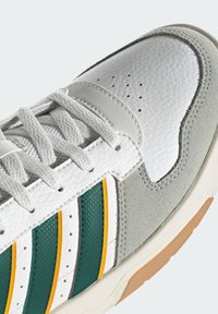 Baskets blanches en suède gris, détails rayés verts et jaunes, lacets blancs et dessus en cuir texturé en gros plan.