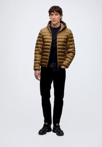 Mann, der eine braune gesteppte Kapuzenjacke, ein schwarzes Hemd, schwarze Hose und schwarze Sneakers trägt, steht vor einem einfarbigen hellen Hintergrund.
