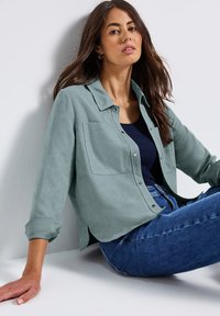 Žena s dlouhými hnědými vlasy sedící opřená o bílou zeď, oblečená v zelené košili na knoflíky, černém topu a modrých džínách.