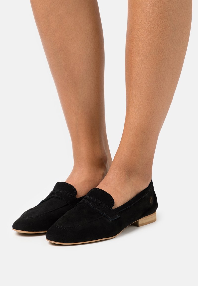 Pieds portant des mocassins à enfiler en daim noir avec des talons bas couleur beige sur un fond blanc.
