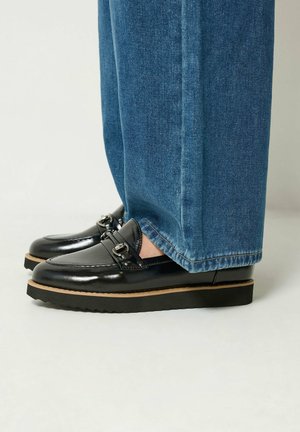 Mocassins en cuir noir avec détails de boucle argentée portés avec un jean bleu sur un fond neutre.