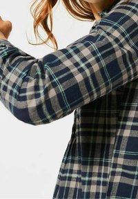 Flannel skjorte med et navy, grå og grøn ternet mønster, lange ærmer og en blød tekstur. Knapper synlige langs sideblinden.