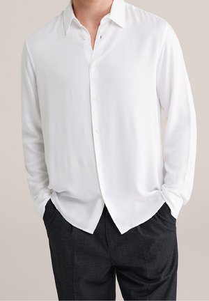 Homme portant une chemise blanche à manches longues boutonnée et un pantalon noir, mains dans les poches, debout devant un fond uni.