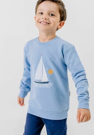 Lächelnder Junge, der ein hellblaues Sweatshirt mit einer weißen Segelboot- und Sonnen-Grafik trägt, kombiniert mit dunkelblauen Hosen.