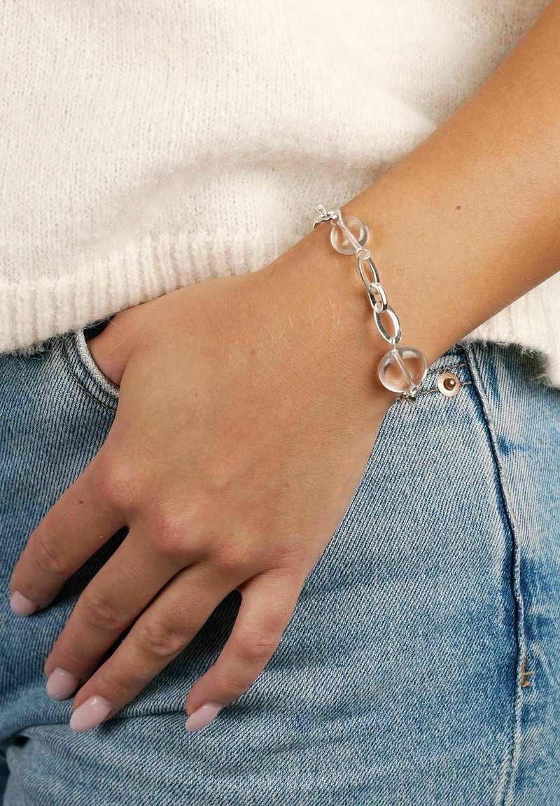 Silverfärgad kedjearmband med cirkulära och ovala länkar, prydd med klara runda pärlor. Bärs på en hand som vilar på denimtyg.