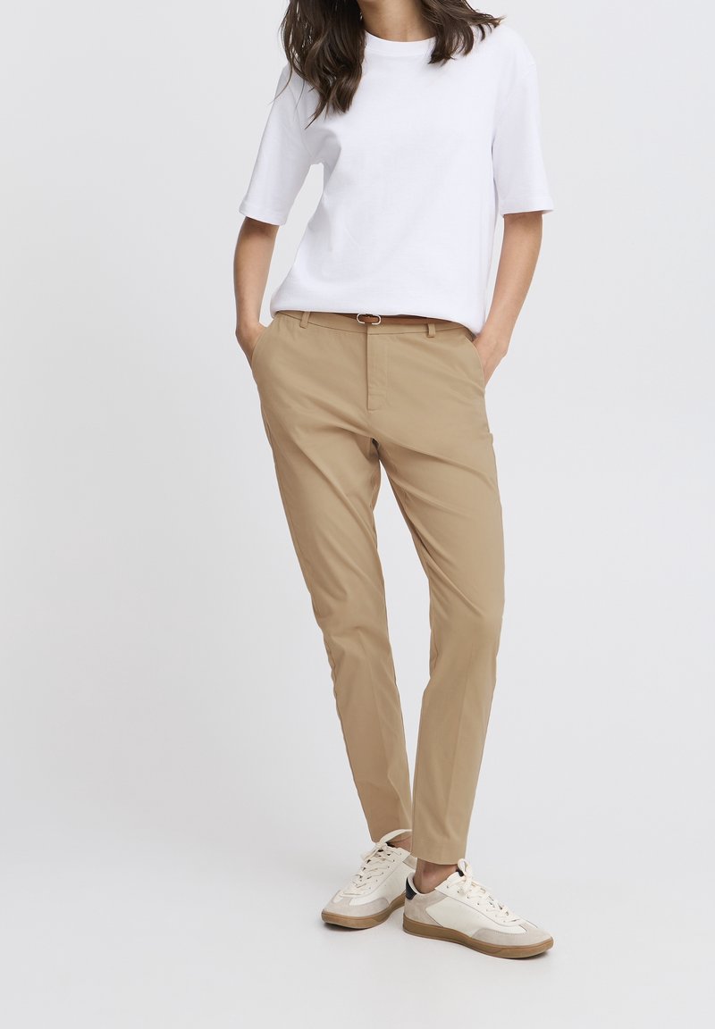 Femme portant un T-shirt blanc, un pantalon beige slim avec ceinture, et des baskets blanches, debout les mains dans les poches devant un fond uni.