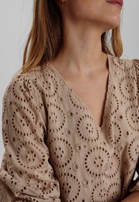 Beige, broderad blus med cirkulära utskurna mönster, V-ringning och puffärmar. Mjukt material med detaljerade stygnaccenter.