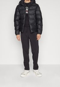 Černá péřová bunda s kapucí, prošívaným designem, předním zipem a elastickými manžetami, v kombinaci s černými kalhotami a bílými teniskami.