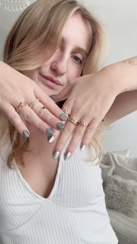 Srebrni metalik lak za nokte na rukama koje prikazuju više nježnih zlatnih prstenova različitih dizajna, na svijetloj pozadini.