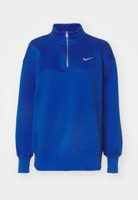 Blå fleece-tröja med hög krage, halv dragkedja och ribbade ärmslut. Har en liten silverlogga på vänster bröst. Mjuk textur.