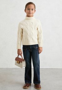 Suéter de punto trenzado crema con cuello alto, jeans azul oscuro con costuras laterales y bolso pequeño beige con acentos marrones y herrajes dorados.