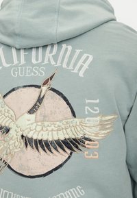 Sudadera de color azul claro con un gran diseño de grúa bordado, con acentos en beige y negro, superpuesto con texto y un detalle circular.