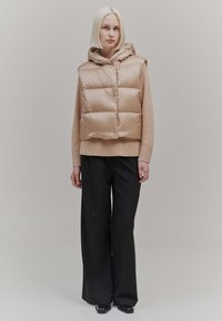 Puffer vest with a hood, beige color, glossy texture, snap buttons, worn over a beige knit sweater, paired with wide black trousers.