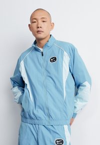 Ljusblå träningsjacka med kontrasterande ljusblå detaljer, framsidan med dragkedja och svart Nike AIR-logotyp på vänster bröst.