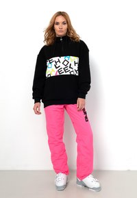 Schwarzer Kapuzenpullover mit buntem Grafikband, kombiniert mit leuchtend rosa Snowboardhosen. Das Modell trägt weiße Schneestiefel mit grauen Akzenten.