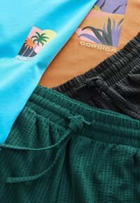 Deux t-shirts graphiques avec des imprimés de palmiers, étiquetés "Nice" et "Corse", associés à des shorts à cordon de serrage texturés verts et noirs.