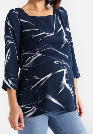 Femme portant un chemisier bleu marine avec des motifs abstraits de coups de pinceau blancs et des manches trois-quarts, associé à un jean bleu clair.