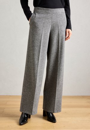 Mujer que lleva pantalones grises de cintura alta y pierna ancha con pliegues delanteros, combinados con botines negros de tacón y un top negro de manga larga.
