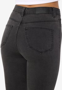 Gros plan d'une personne portant un jean en denim noir ajusté, montrant les poches arrière et une étiquette de marque "Noisy May" sur la ceinture.