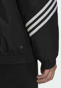 Chaqueta tipo windbreaker negra con rayas blancas en las mangas, puños y dobladillo elásticos, y un pequeño detalle de logo cerca de la parte inferior.